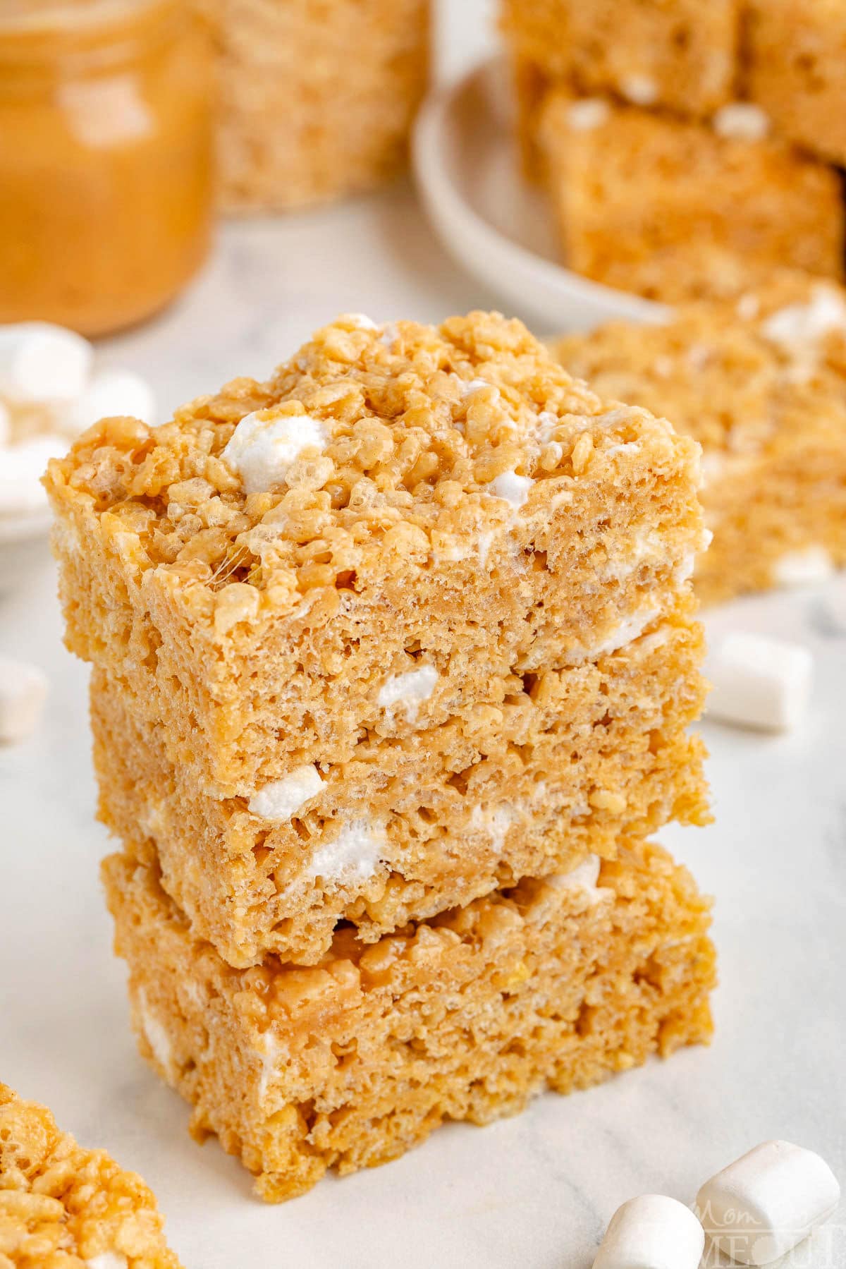 Peanut butter rice krispie treats stacked three high on white surface with marshmallows scattered about.