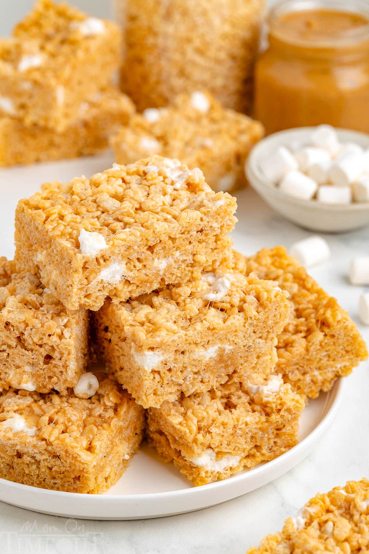 Peanut butter rice krispie treats stacked on a white round plate with more treats in the background.