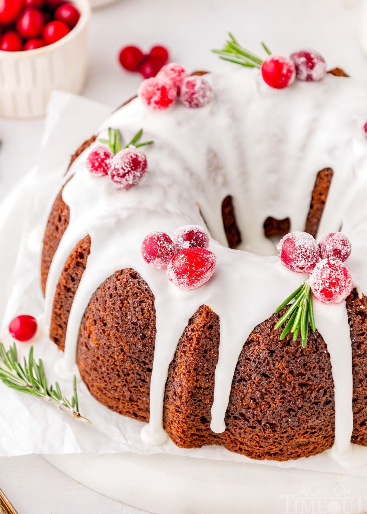 Gingerbread bundt cake topped with vanilla icing and cranberry and rosemary garnishes. The cake is on a white cake stand.