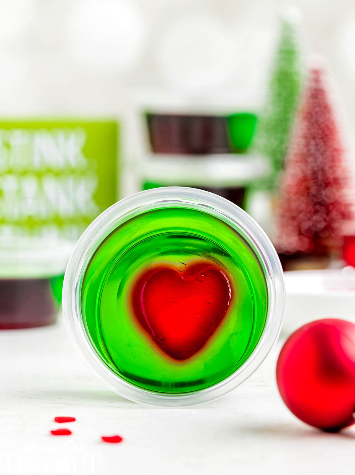 A green and red grinch jello shot in a shot glass laying on its side. Christmas decorations are in the background.