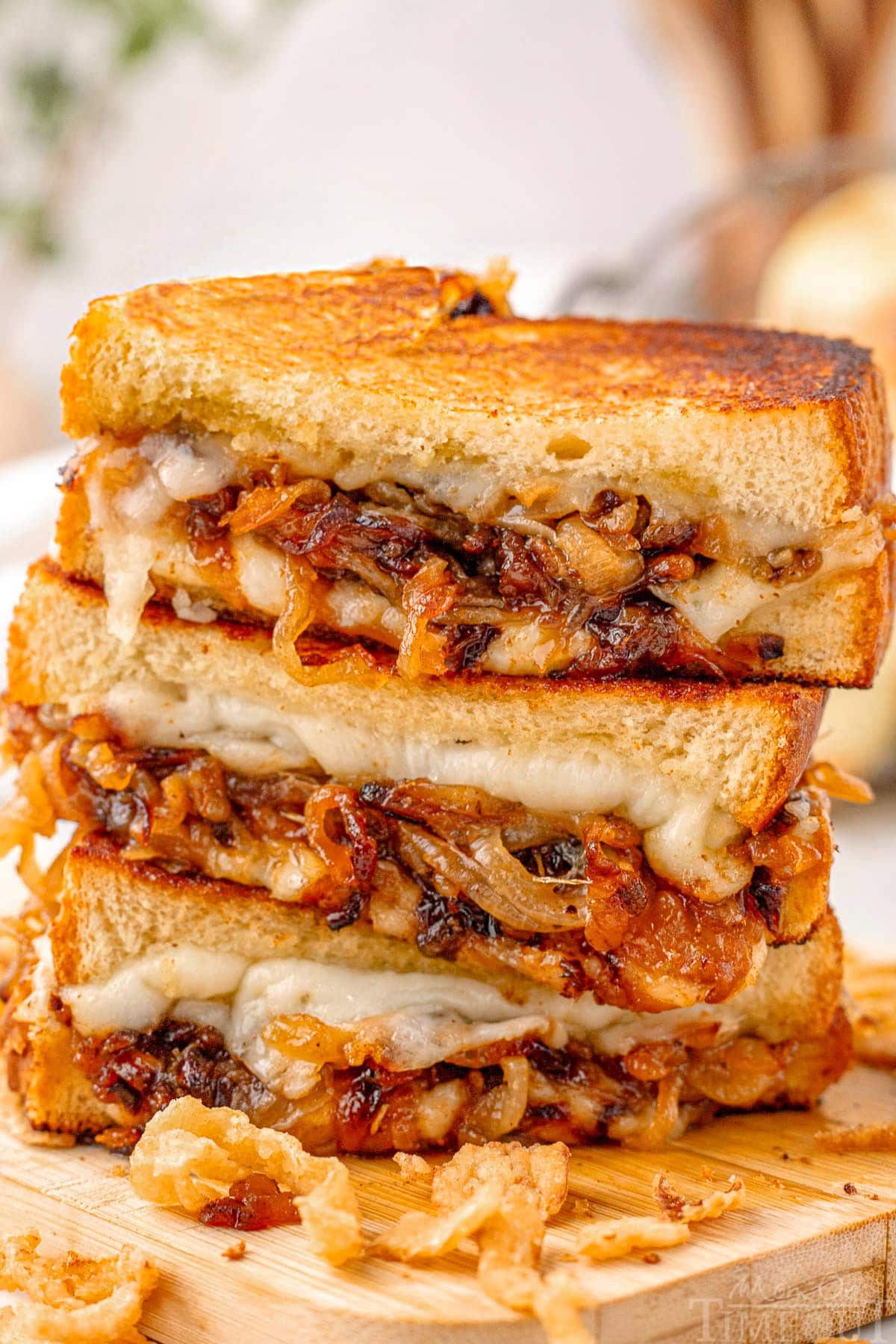 Three halves of a french onion grilled cheese sandwich stacked on a wood cutting board with french fried onions surrounding it.