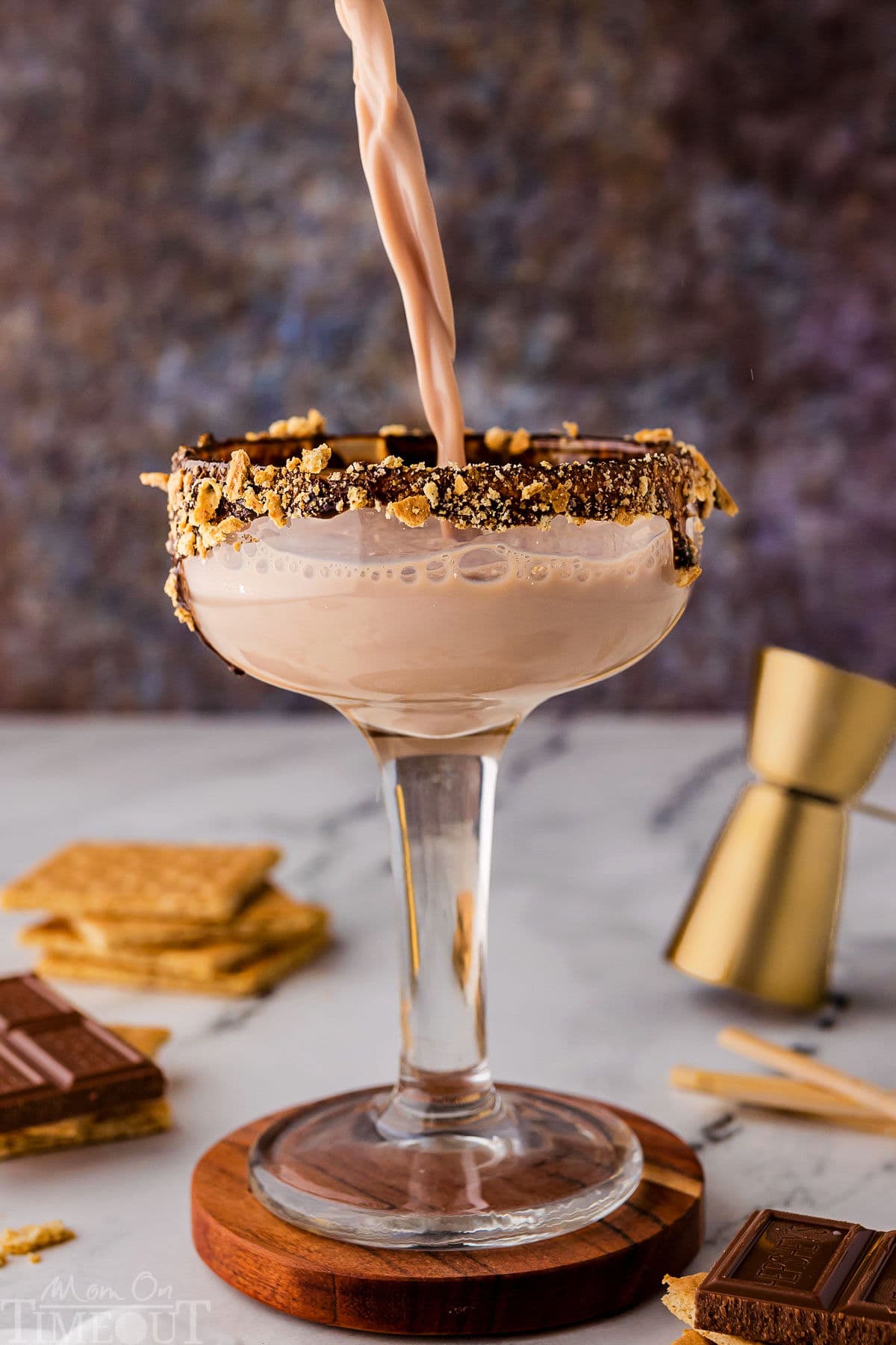 Pouring smores martini into the glass that's on top of a wood coaster. Recipe ingredients are sitting on the surface nearby.