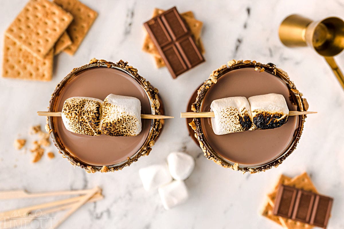 A top down view of two martini glasses with smores martini topped with toasted marshmallows. Recipe ingredients are scattered on the surface.
