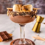Side view of smores martini in a tall glass sitting on a wood coaster. Recipe ingredients are laying on the surface nearby.
