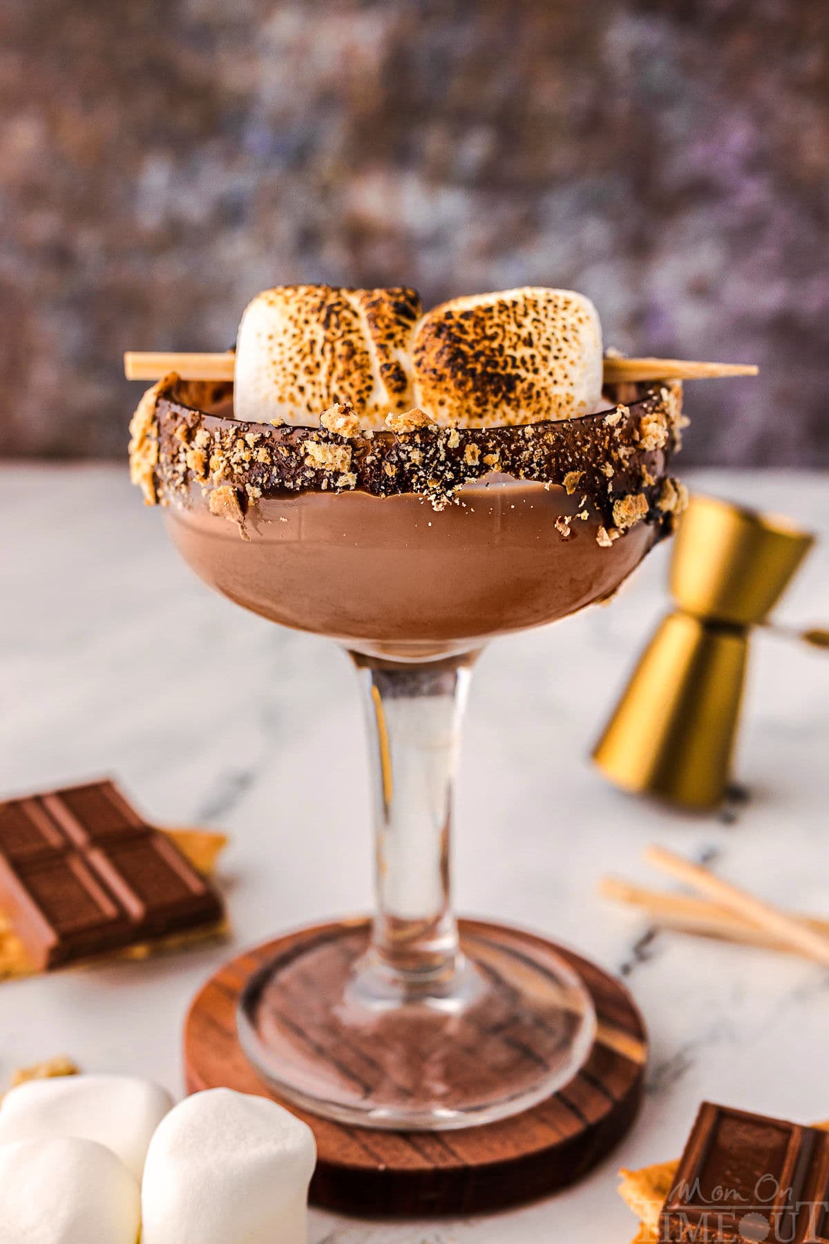 Side view of smores martini in a tall glass sitting on a wood coaster. Recipe ingredients are laying on the surface nearby.