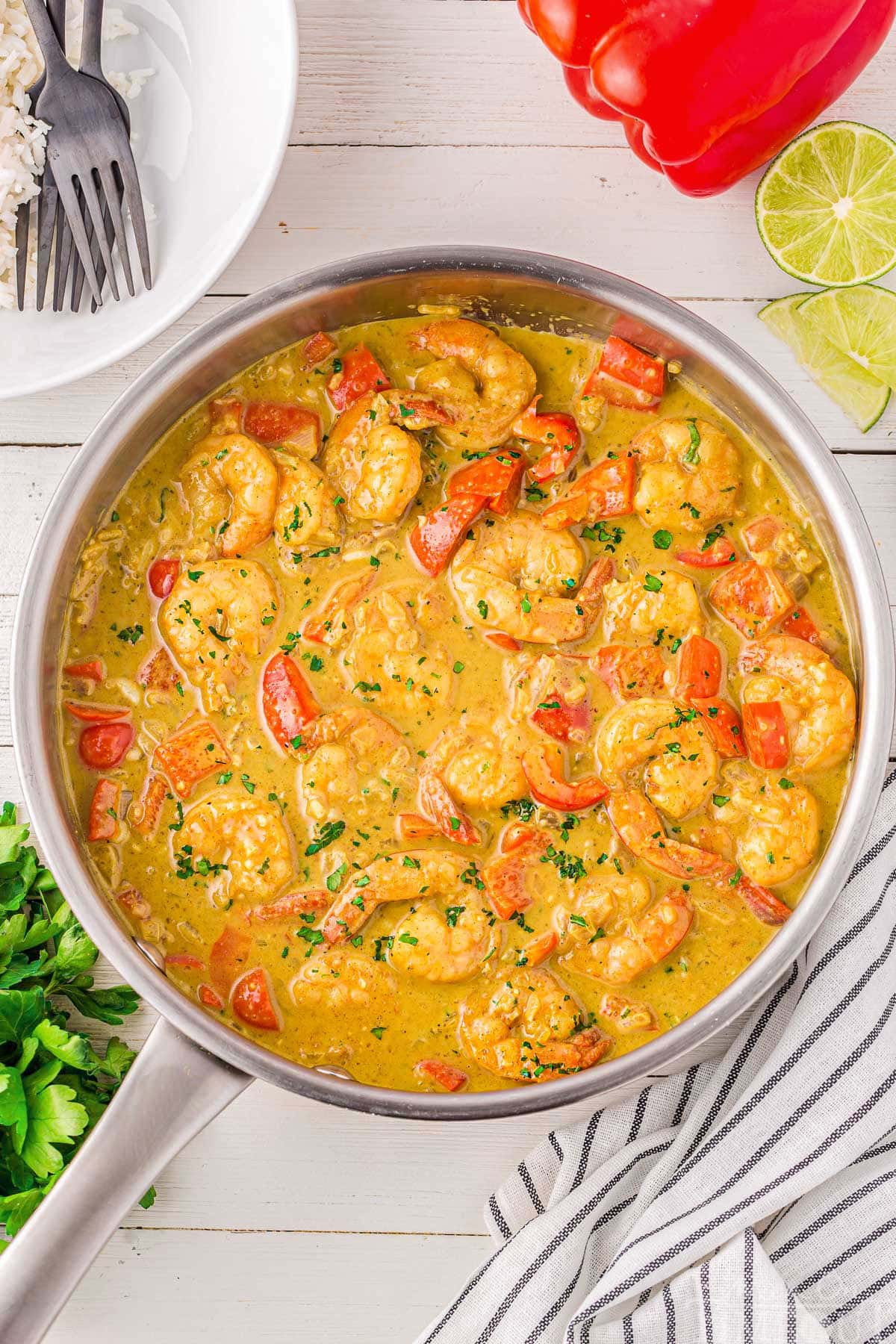 Shrimp Curry in a metal skillet. Ingredients are placed around the skillet, as well as plates and forks and a striped linen.