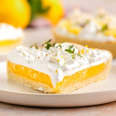 A side view of a slice of lemon meringue tart on a white plate. Extra slices of the tart and lemons are in the background.