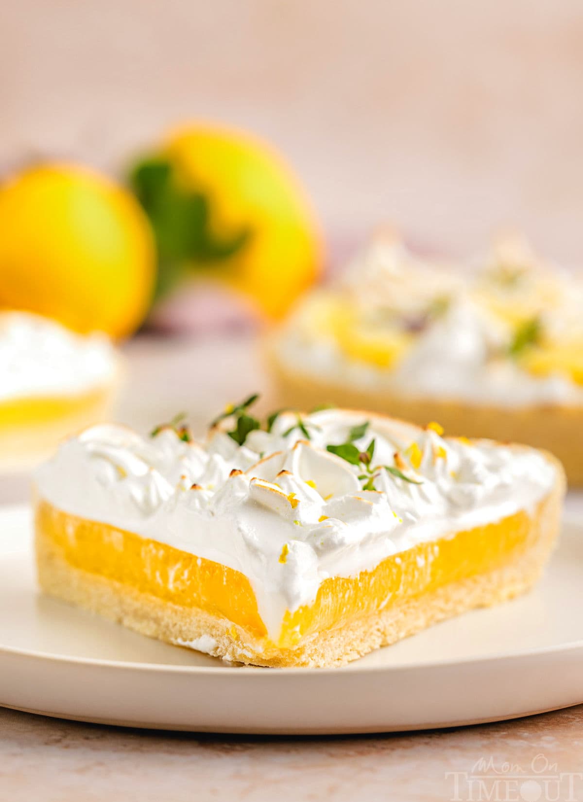 A side view of a slice of lemon meringue tart on a white plate. Extra slices of the tart and lemons are in the background.