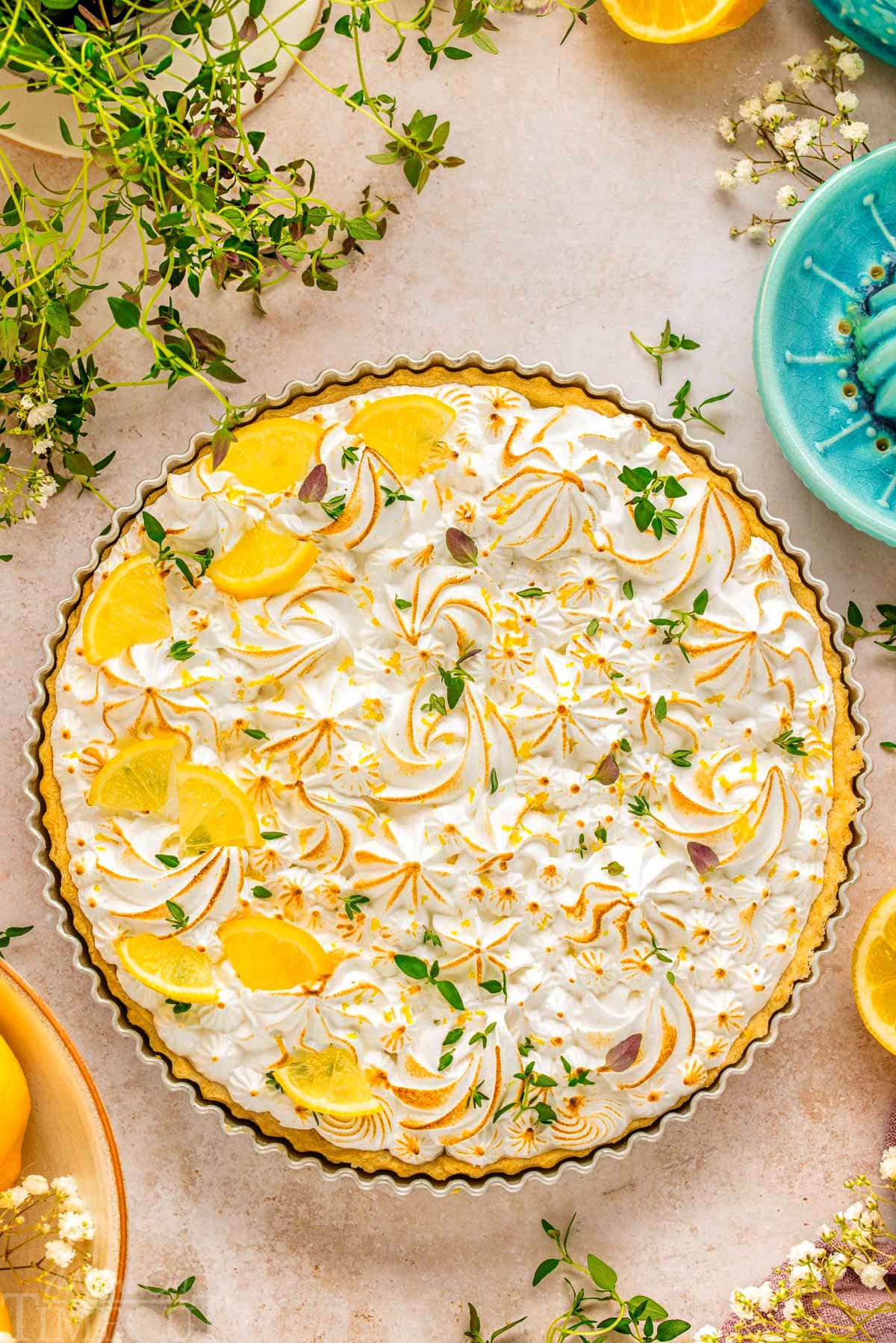 A top down view of Lemon Meringue Tart. The tart is topped with lemon wedges and chopped lemon thyme.