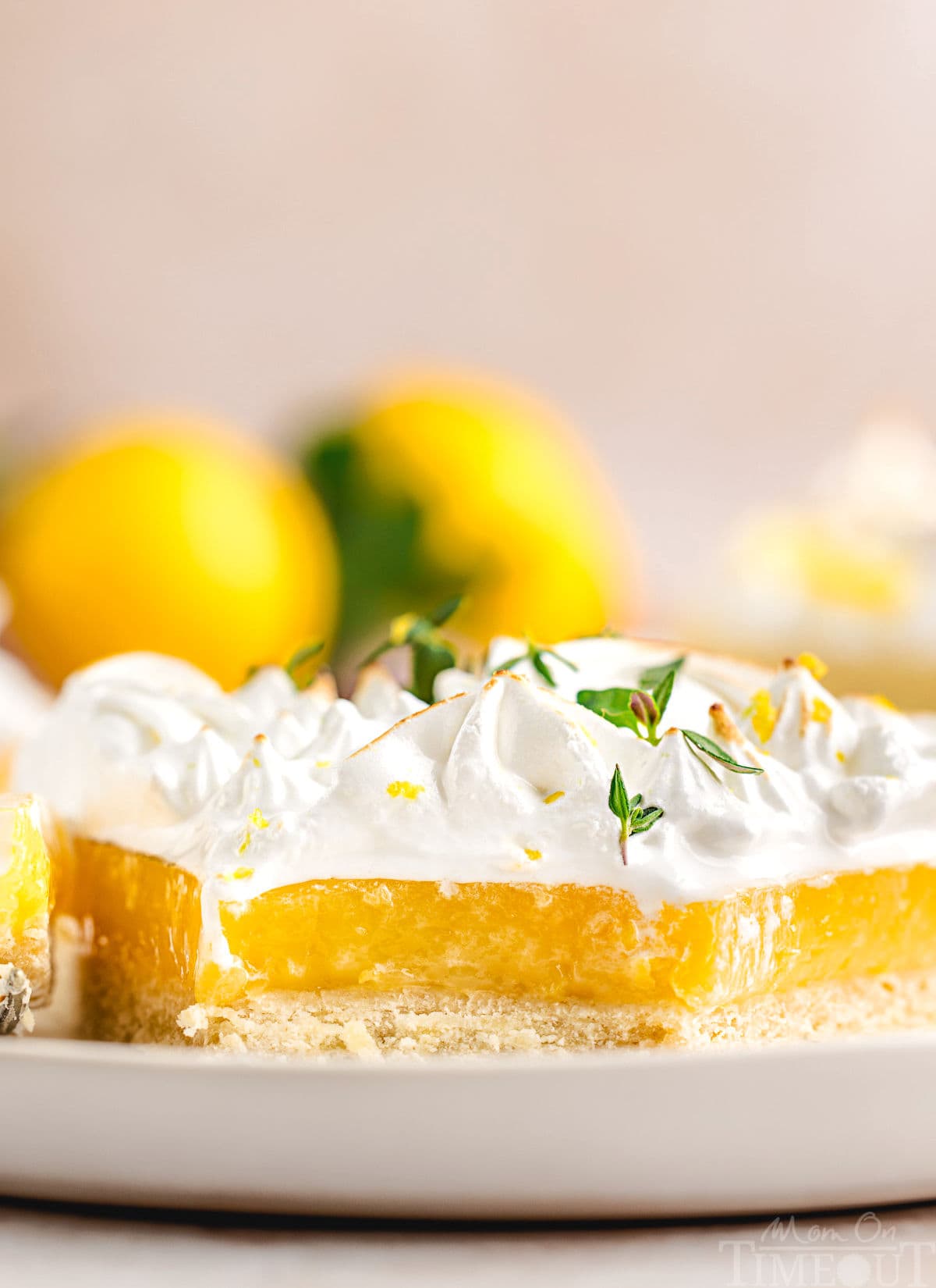 A side view of a slice of lemon meringue tart with a bite taken out of it. Lemons are in the background.