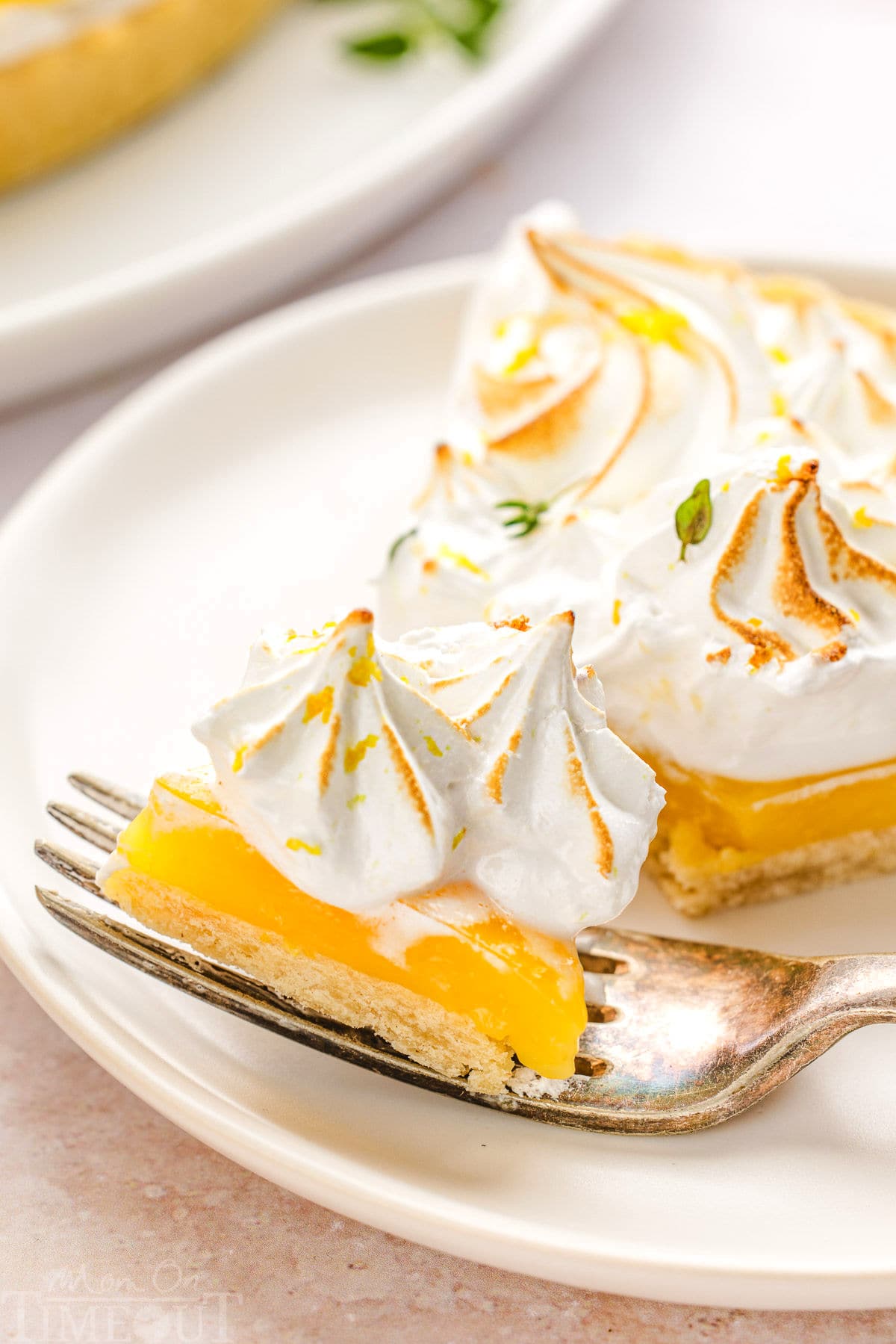 A close up view of a slice of lemon meringue tart on a white plate. A fork is holding a bite of the tart.