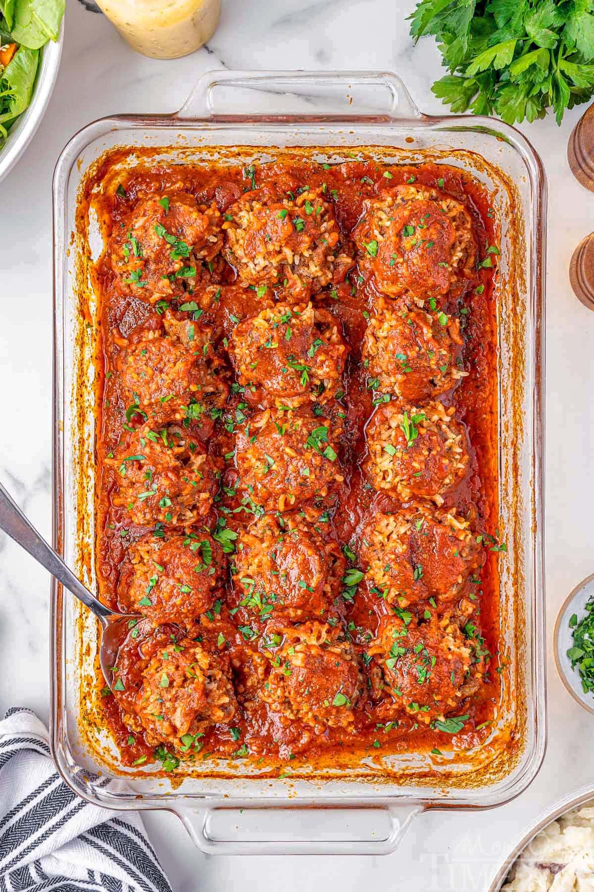 Porcupine Meatballs with fresh parsley sprinkled on top. The meatballs are lined up in a baking pan. A metal spoon is holding one of the meatballs. Ingredients are placed around the pan.