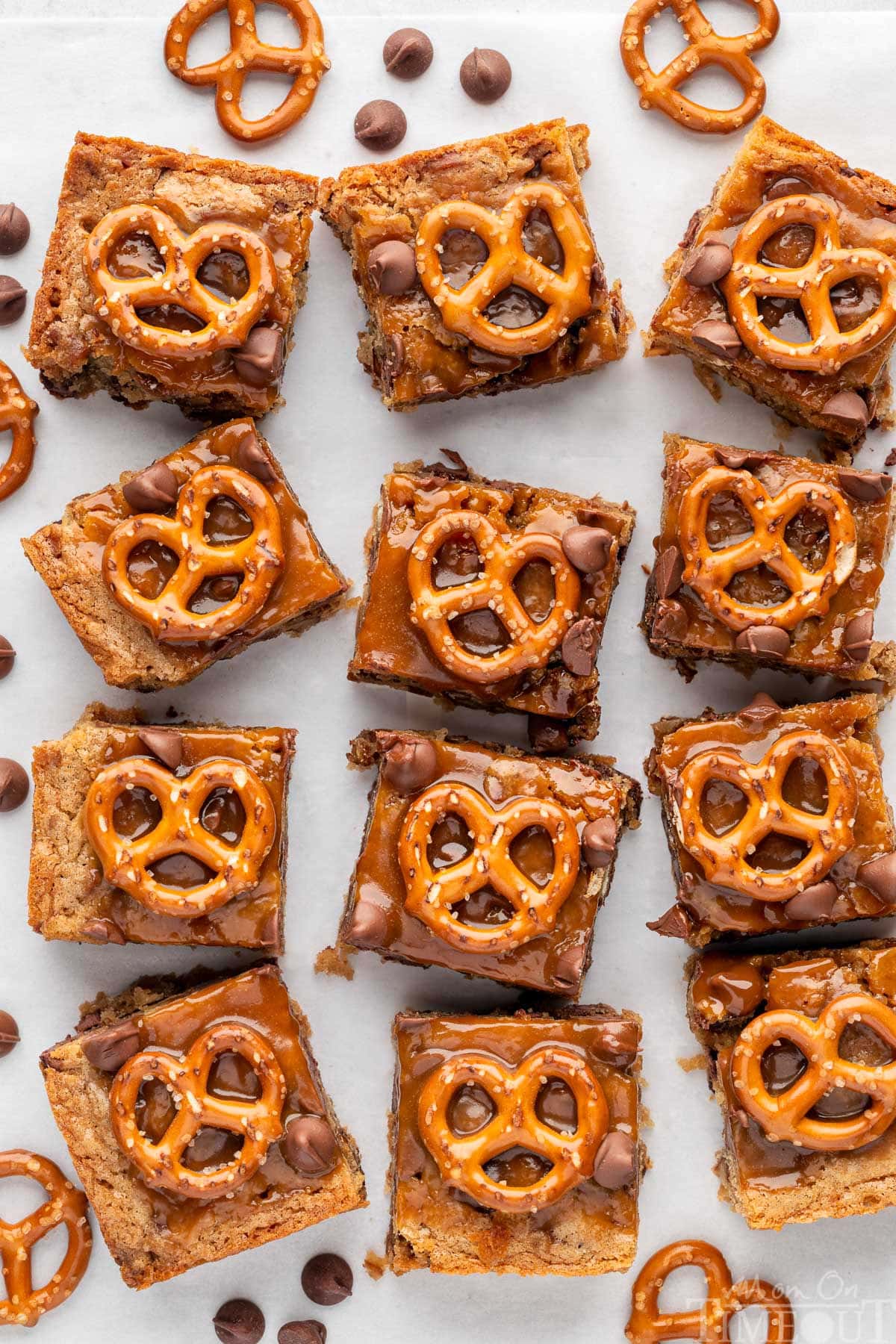Salted Caramel Pretzel Blondies lined up on a piece of parchment paper. Pretzels and chocolate chips are scattered around the bars.