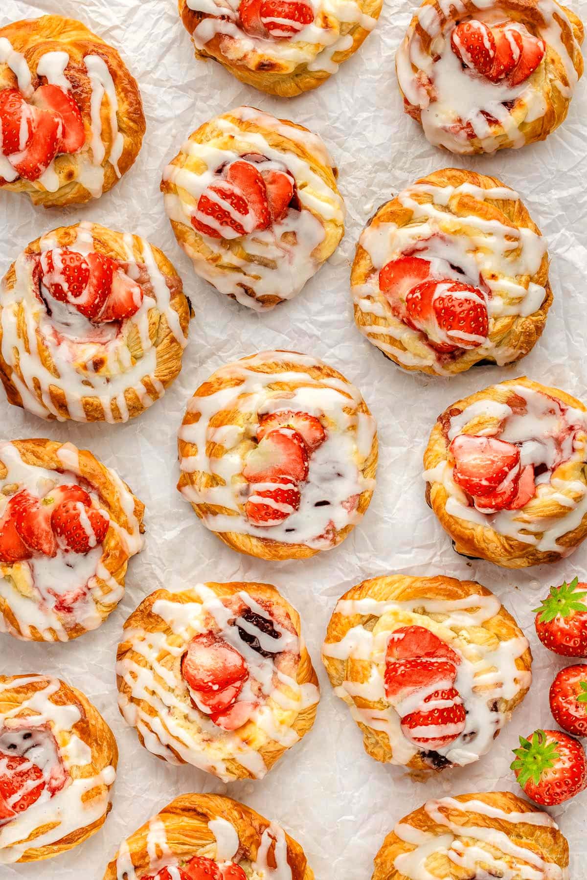 Top down look at strawberry cream cheese danishes on parchment paper. About 15 strawberry danishes are shown.
