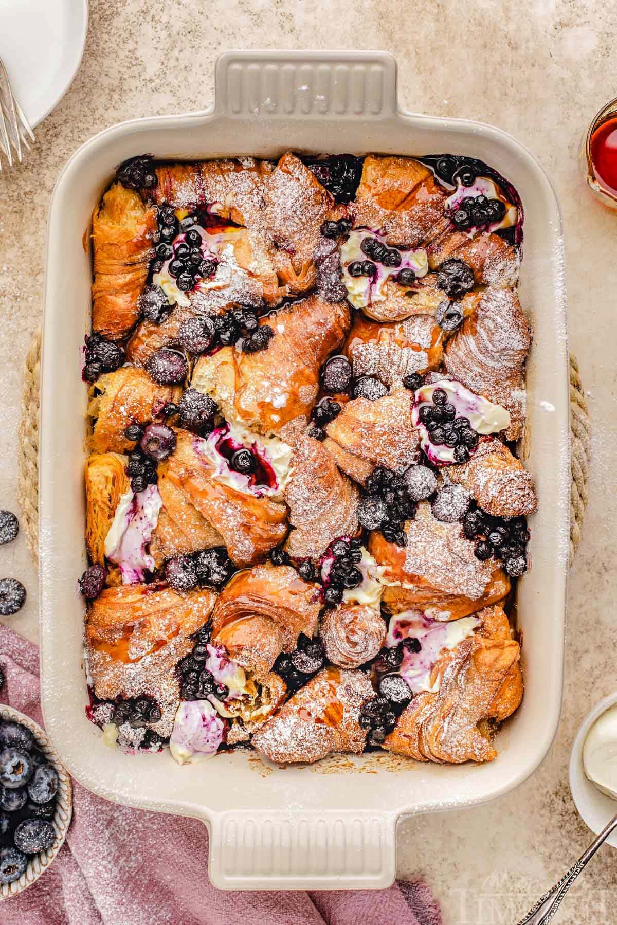 Top down look at croissant french toast bake in baking dish made with blueberries and topped with a dusting of powdered sugar.