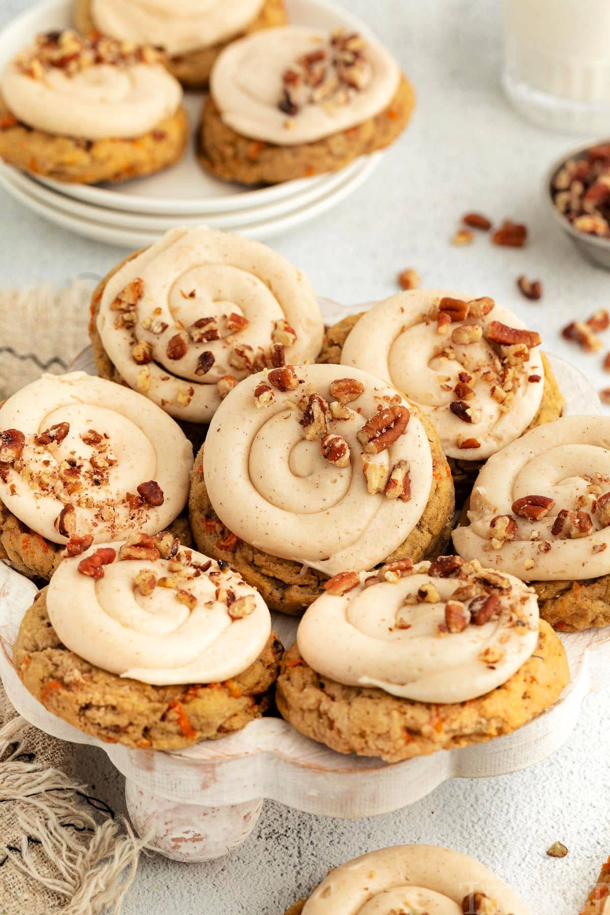 Carrot Cake Cookies placed on a white wooden cake stand. More cookies are in the background on a white plate, and pecans are scattered around.