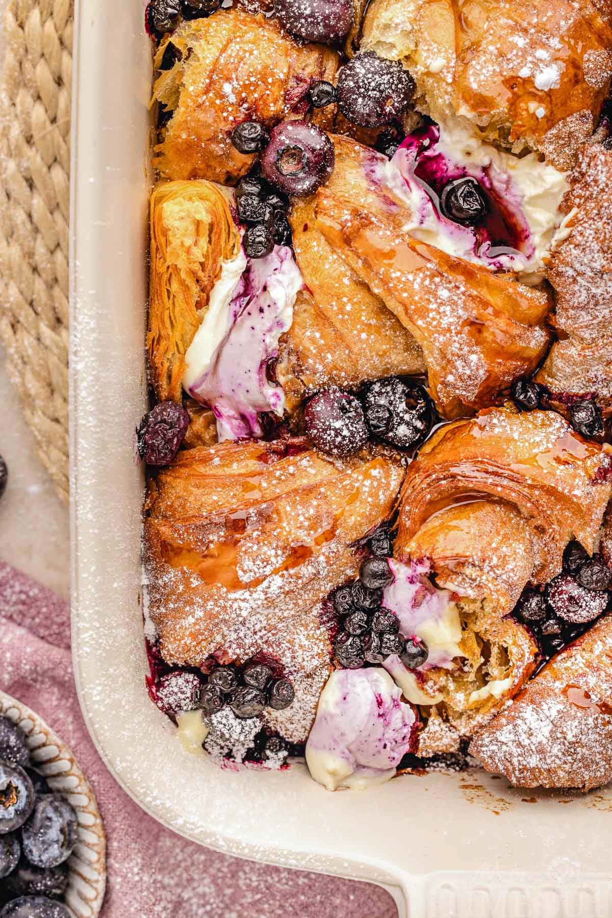 Top down look at croissant french toast bake in baking dish made with blueberries and topped with a dusting of powdered sugar.