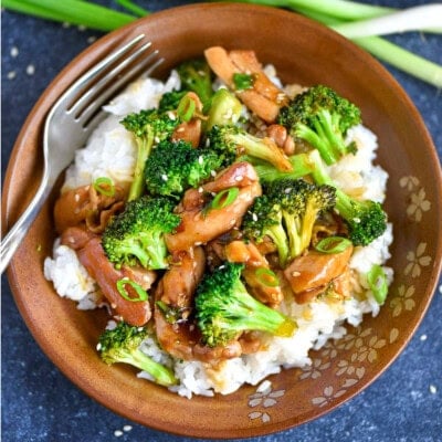 Top down shot of sesame chicken with broccoli recipe in a clay bowl with a bed of white rice. Green onions are to the side of the bowl.