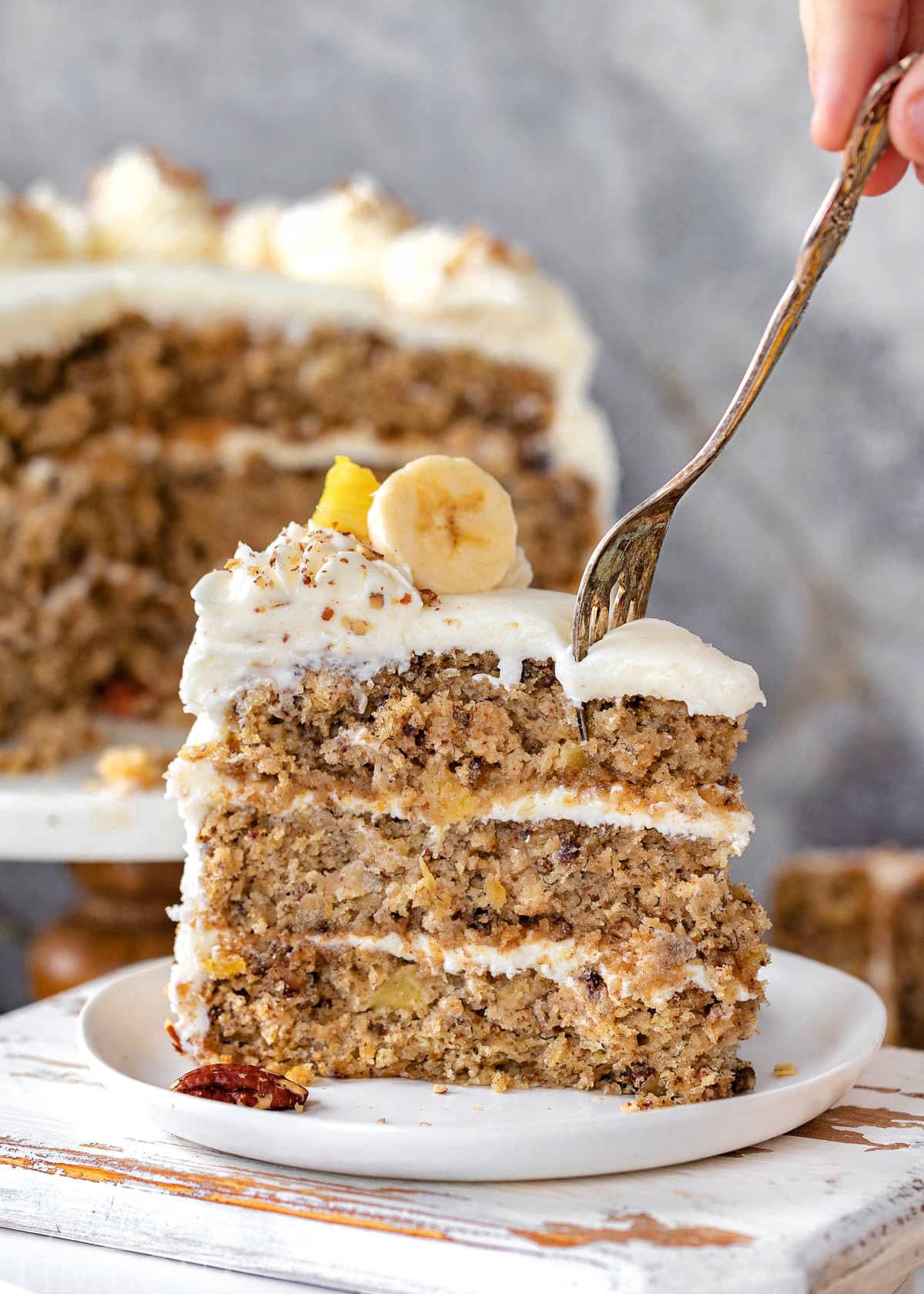Slice of hummingbird cake topped with banana and pineapple and made with cream cheese frosting. Fork cutting into the top of the piece of cake. Rest of cake in background.