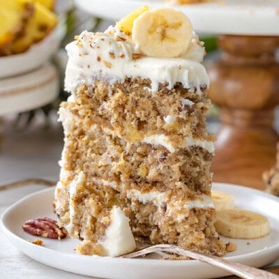Slice of hummingbird cake recipe on a white round plate sitting in front of cake stand with rest of cake on it.