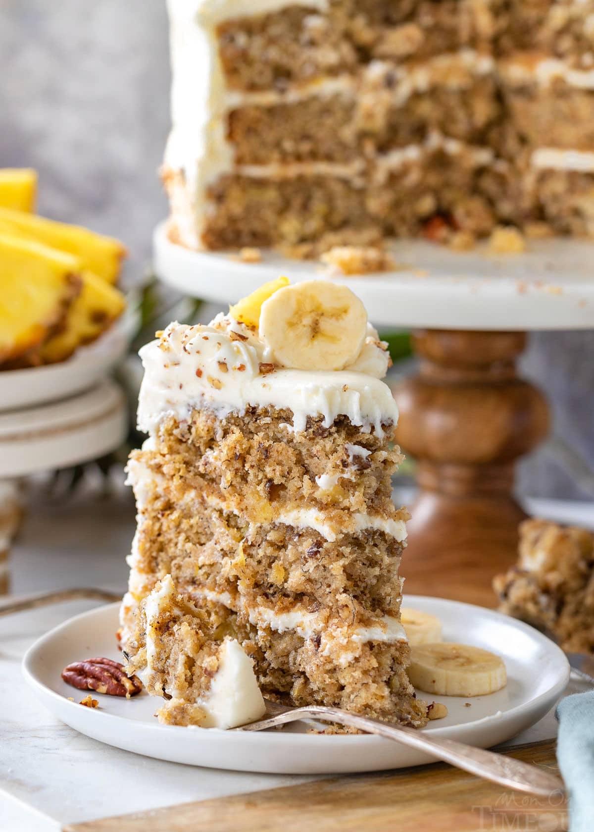 Slice of hummingbird cake recipe on a white round plate sitting in front of cake stand with rest of cake on it.