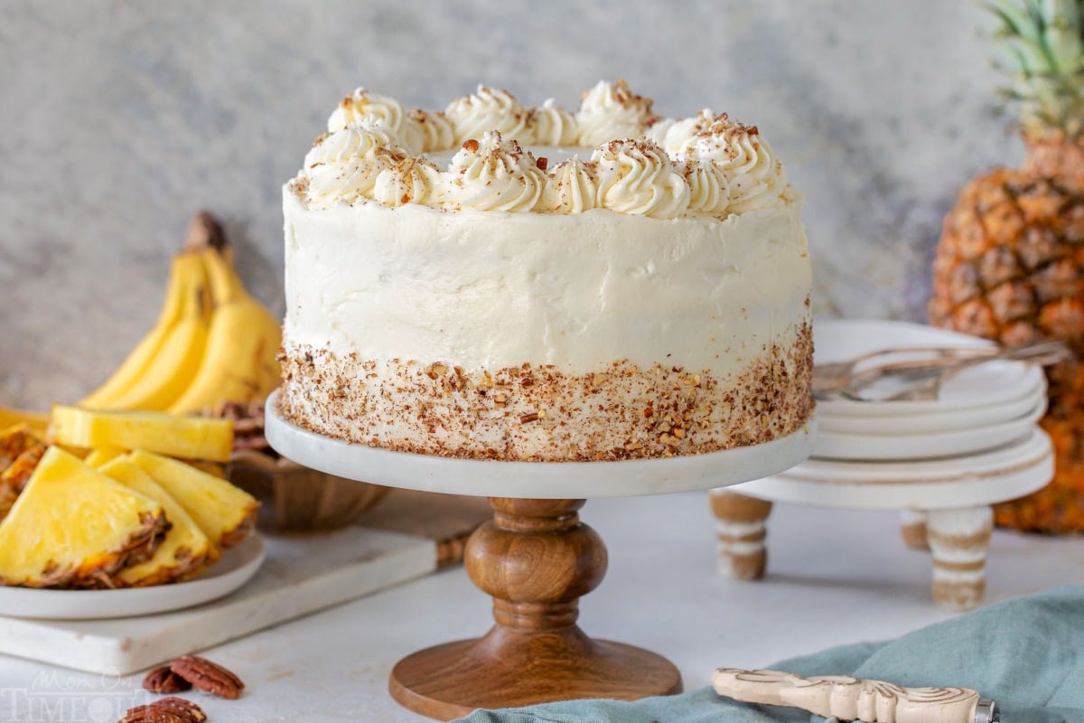 Whole Hummingbird Cake on marble cake stand in front of fresh bananas and pineapple. Small plates can be seen in the background.