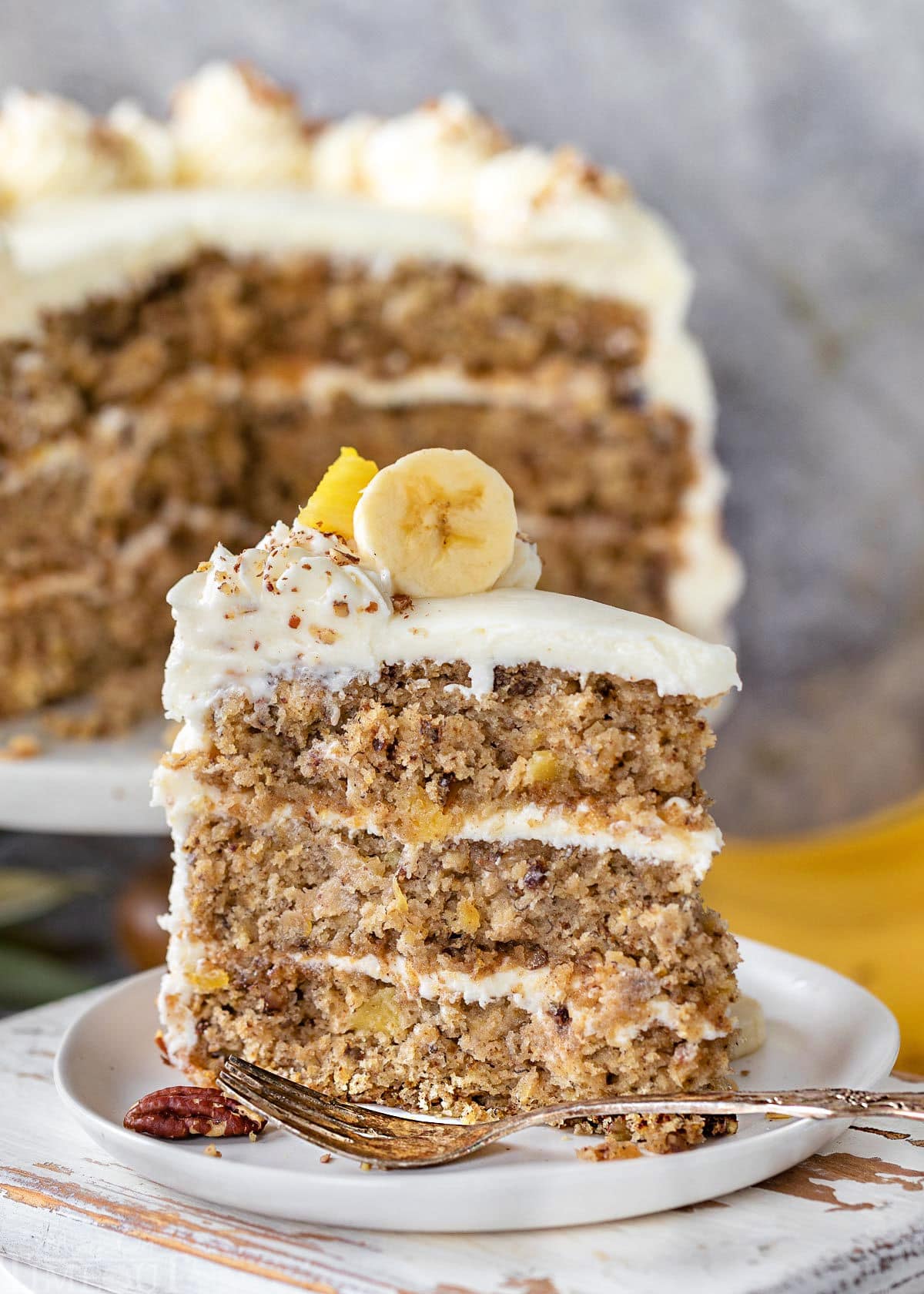 Slice of hummingbird cake topped with banana and pineapple and made with cream cheese frosting. Rest of cake in background.