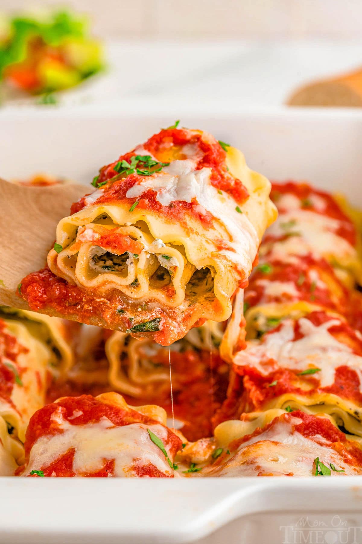 Lasagna roll up being lifted from baking dish with a wood spoon.