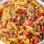 Big serving bowl of Cowboy Pasta Salad ready to be served. A plate of the salad can be seen in the background along with a few limes.