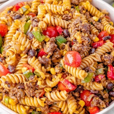 Big serving bowl of Cowboy Pasta Salad ready to be served. A plate of the salad can be seen in the background along with a few limes.