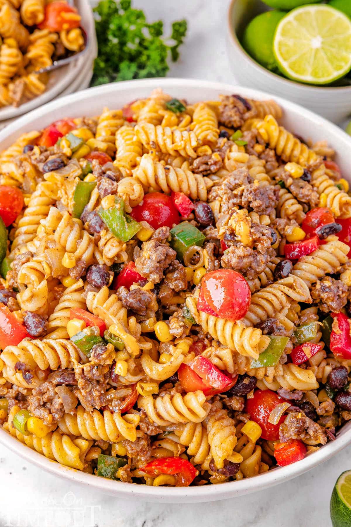 Big serving bowl of Cowboy Pasta Salad ready to be served. A plate of the salad can be seen in the background along with a few limes.
