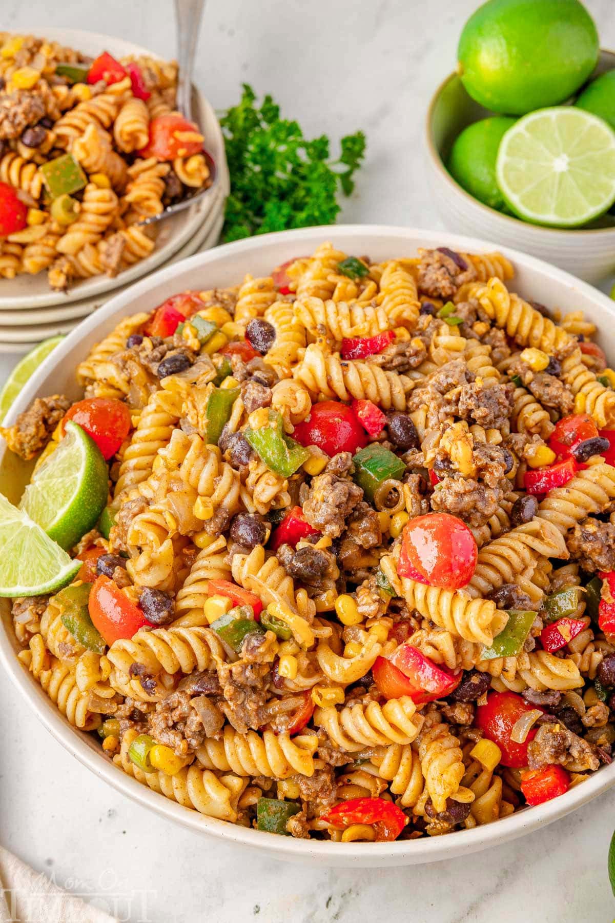 Cowboy pasta salad in white serving bowl garnished with lime wedges. Stack of plates topped with salad in background.