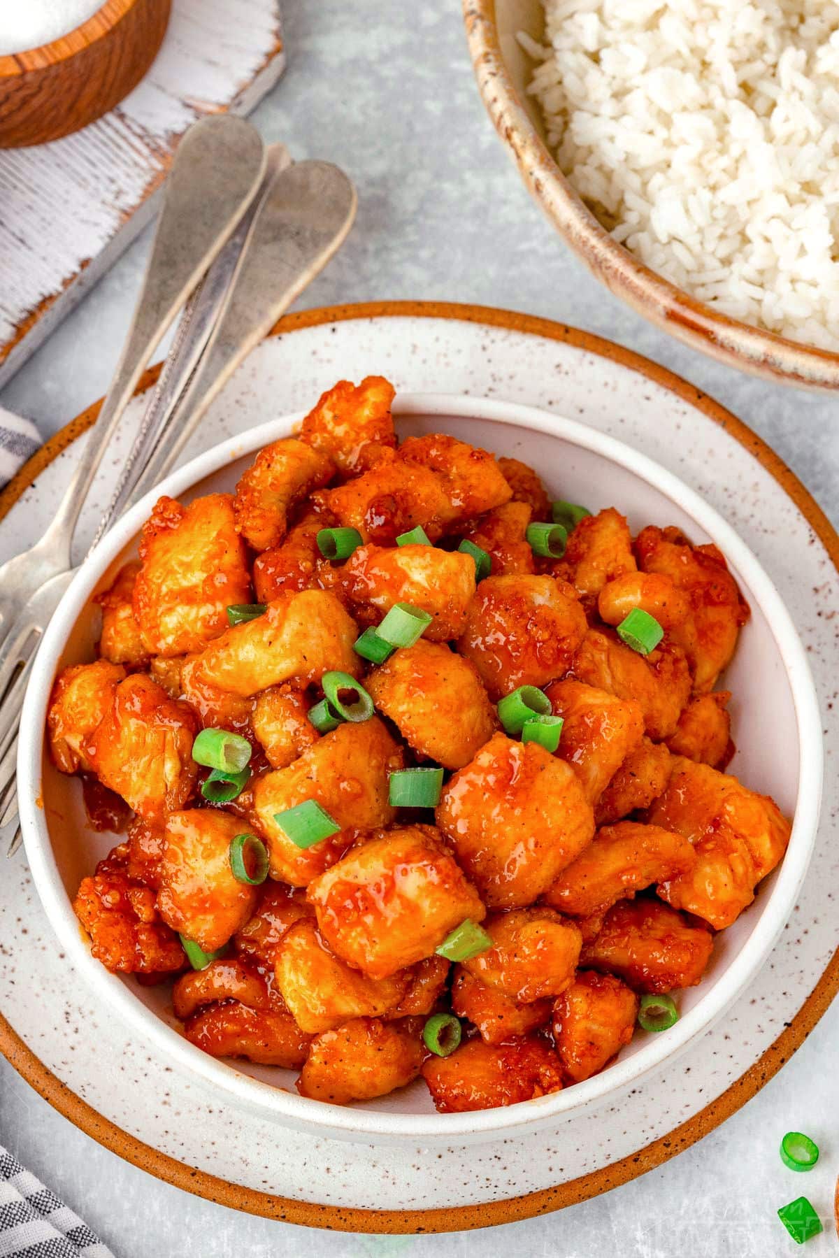 White bowl filled with firecracker chicken and bowl of rice in the background. Chicken is topped with green onions.