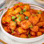White bowl filled with firecracker chicken and bowl of rice in the background. Chicken is topped with green onions.
