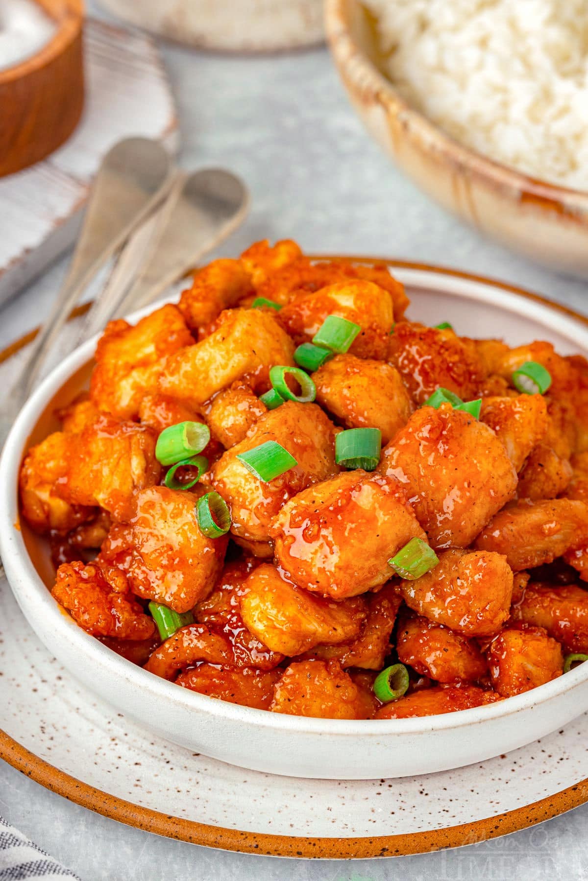 White bowl filled with firecracker chicken and bowl of rice in the background. Chicken is topped with green onions.