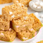 Peanut butter rice krispie treats stacked on a white round plate with more treats in the background.