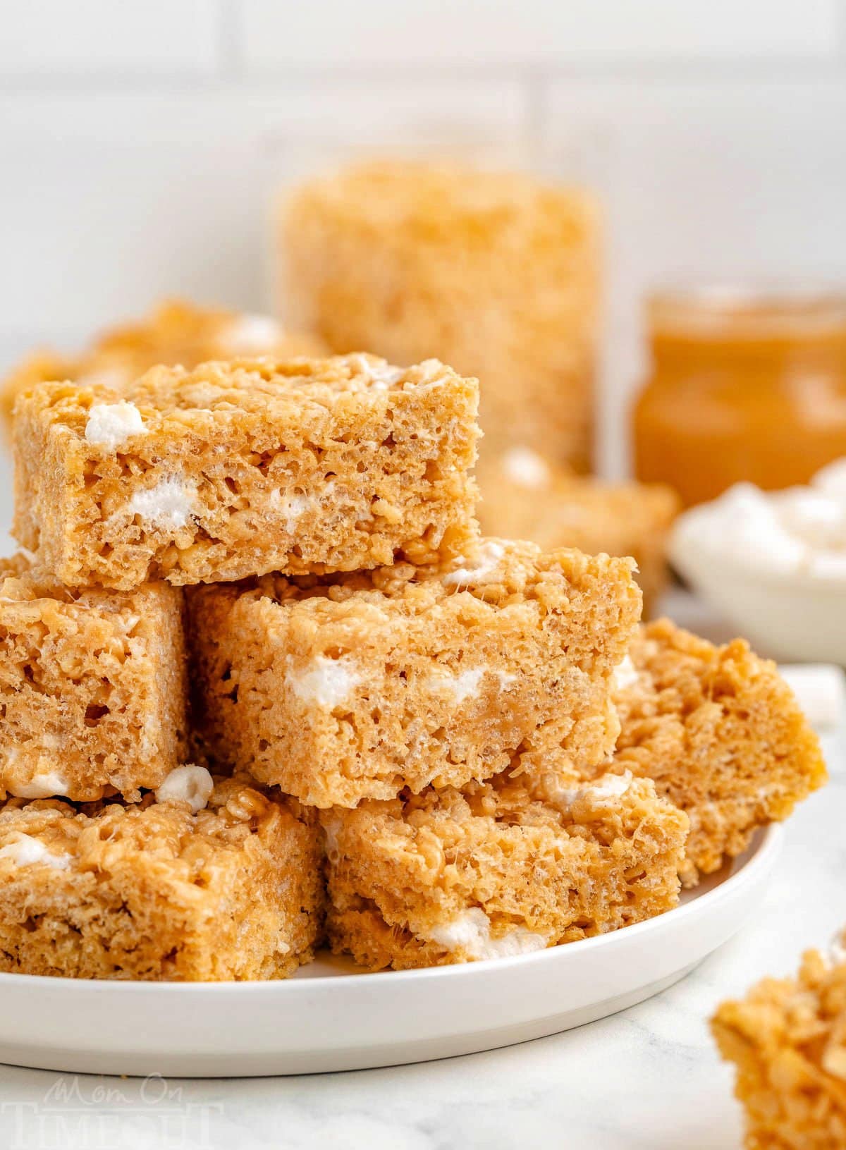 Peanut butter rice krispie treats stacked on a white round plate with more treats in the background.