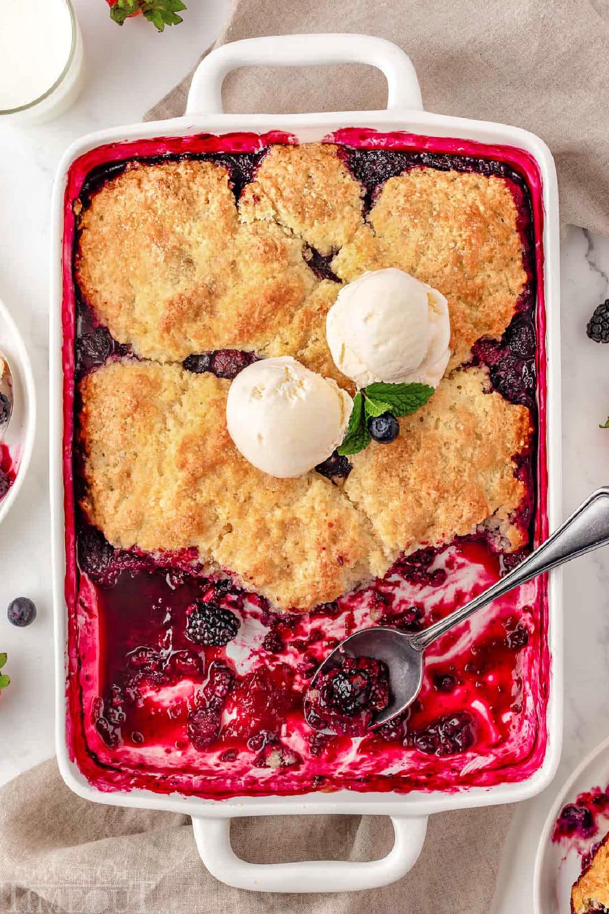 Top down shot of berry cobbler recipe in white baking dish with some servings of the cobbler removed. Topped with vanilla ice cream and fresh mint leaves.