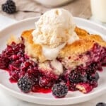 Berry cobbler served on white round plate and topped with vanilla ice cream. Baking dish with the rest of the berry cobbler recipe can be seen in the background.