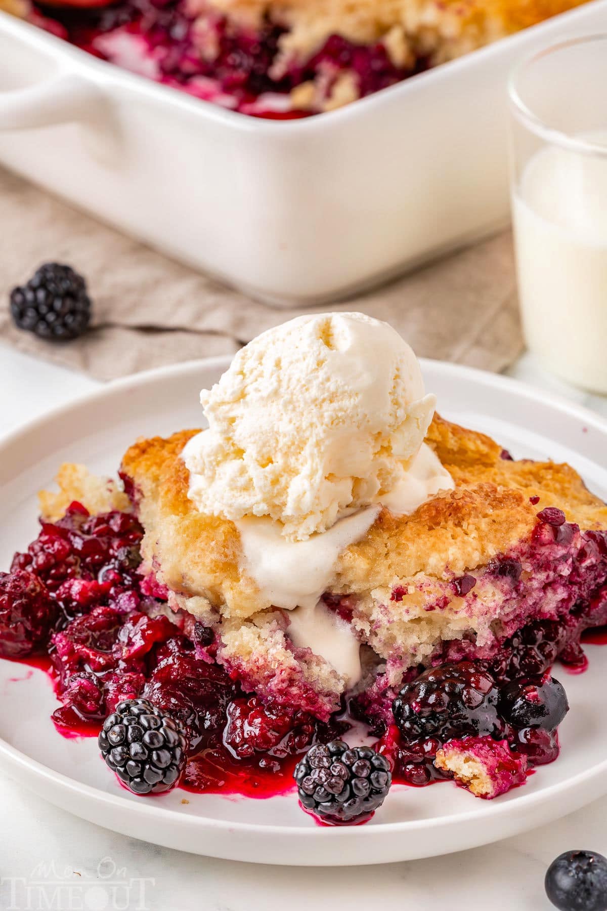 Berry cobbler served on white round plate and topped with vanilla ice cream. Baking dish with the rest of the berry cobbler recipe can be seen in the background.