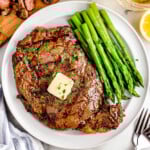 Grilled ribeye steak recipe plated with asparagus on a white round plate next to wood cutting board with sliced grilled steak on it.