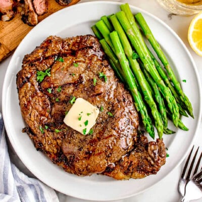 Grilled ribeye steak recipe plated with asparagus on a white round plate next to wood cutting board with sliced grilled steak on it.