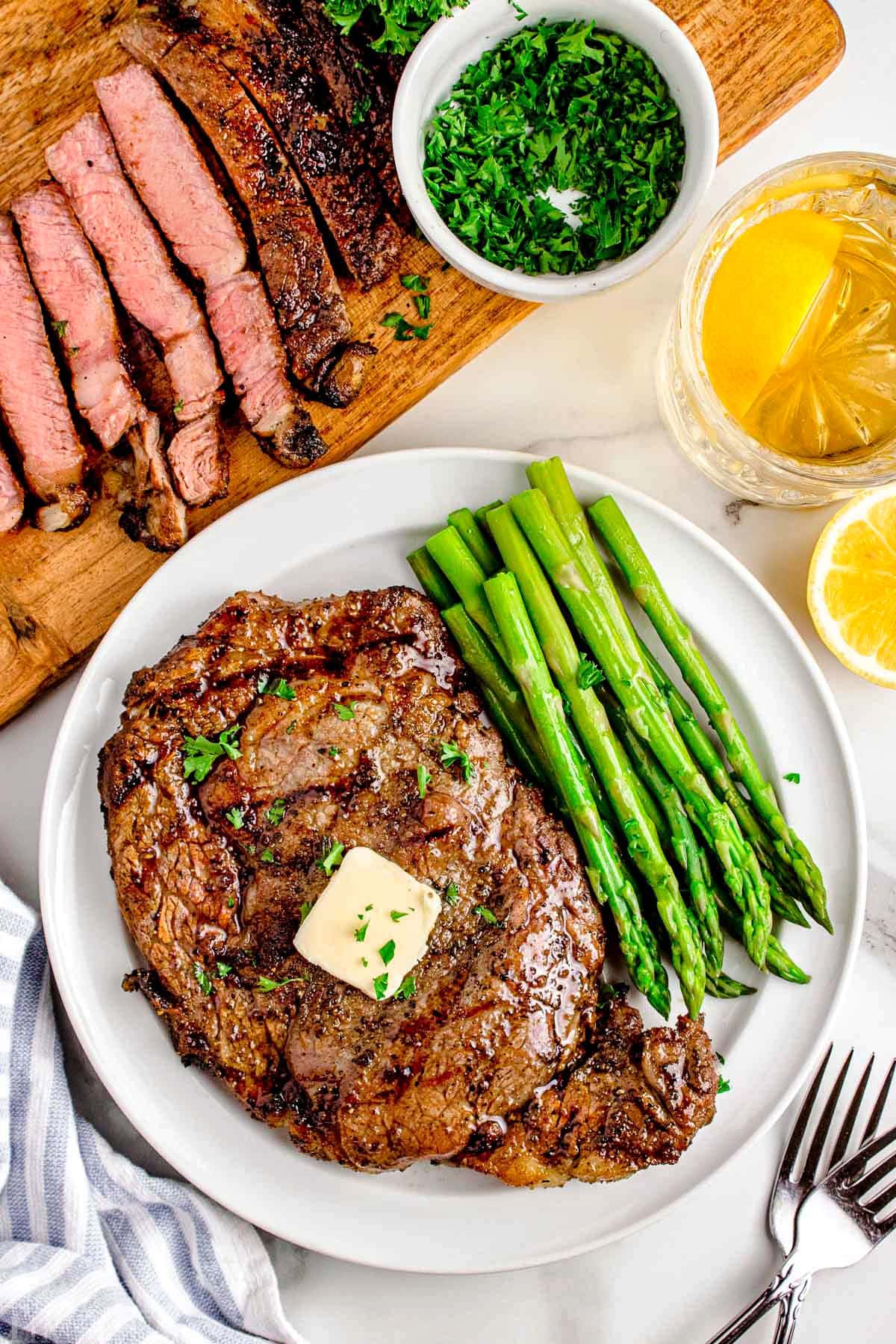 Grilled ribeye steak recipe plated with asparagus on a white round plate next to wood cutting board with sliced ribeye steak on it.