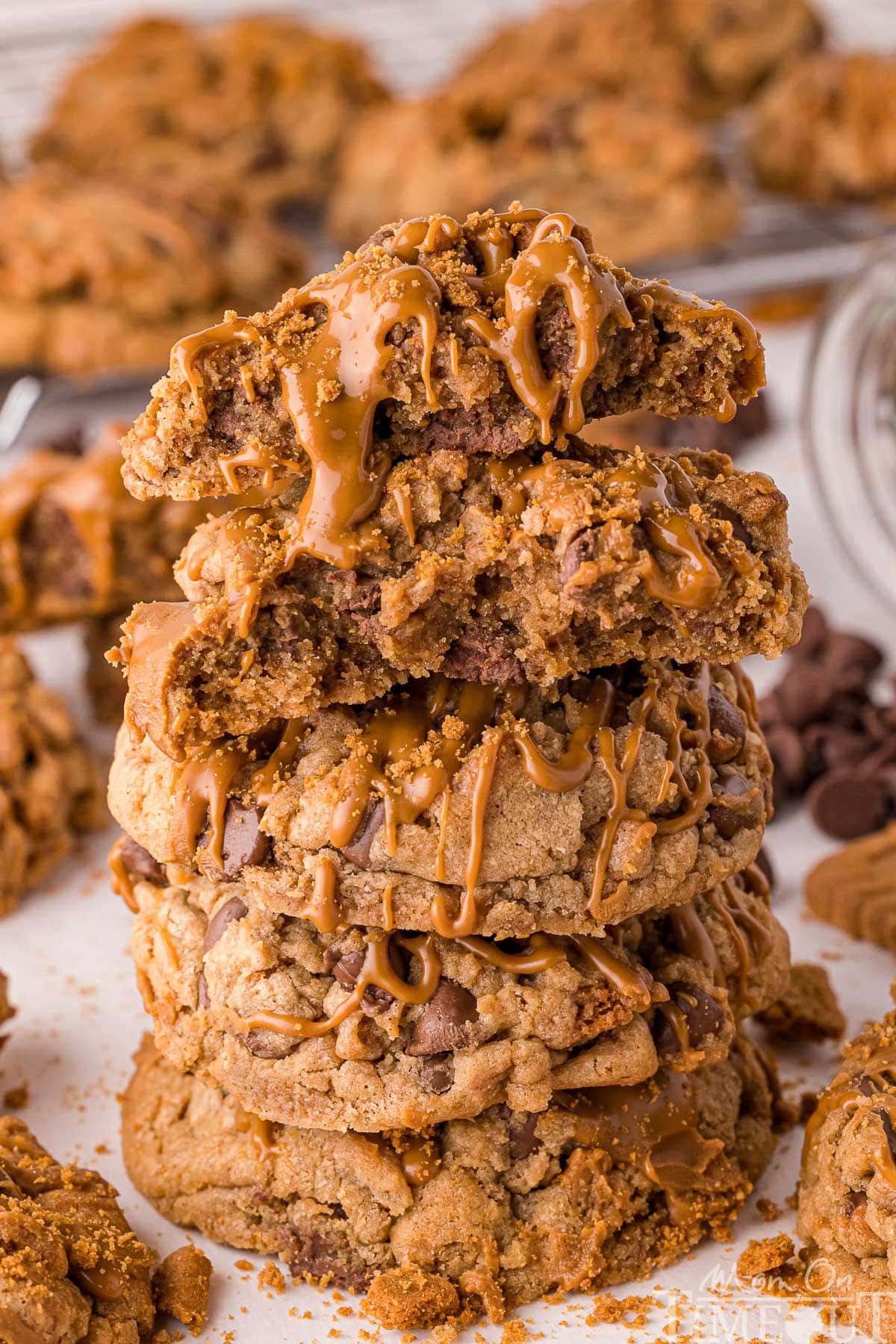Stack of four biscoff chocolate chip cookies topped with cookie butter stacked on white parchment. Top cookie is broken in half. More cookies can be seen in the background.