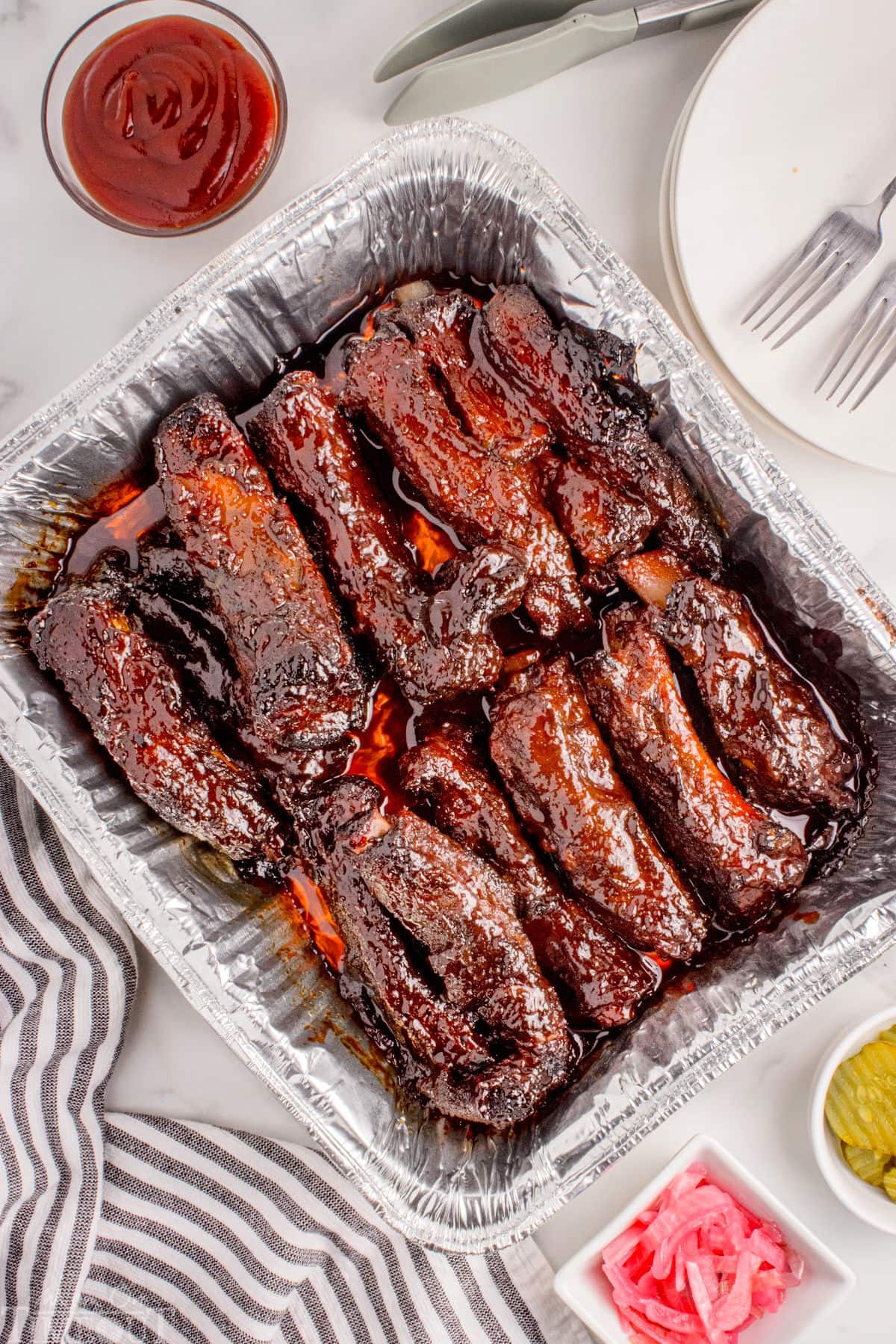 Overhead shot of grilled party ribs in foil pan with bbq sauce, pickled onions and pickles in small bowls to the side.