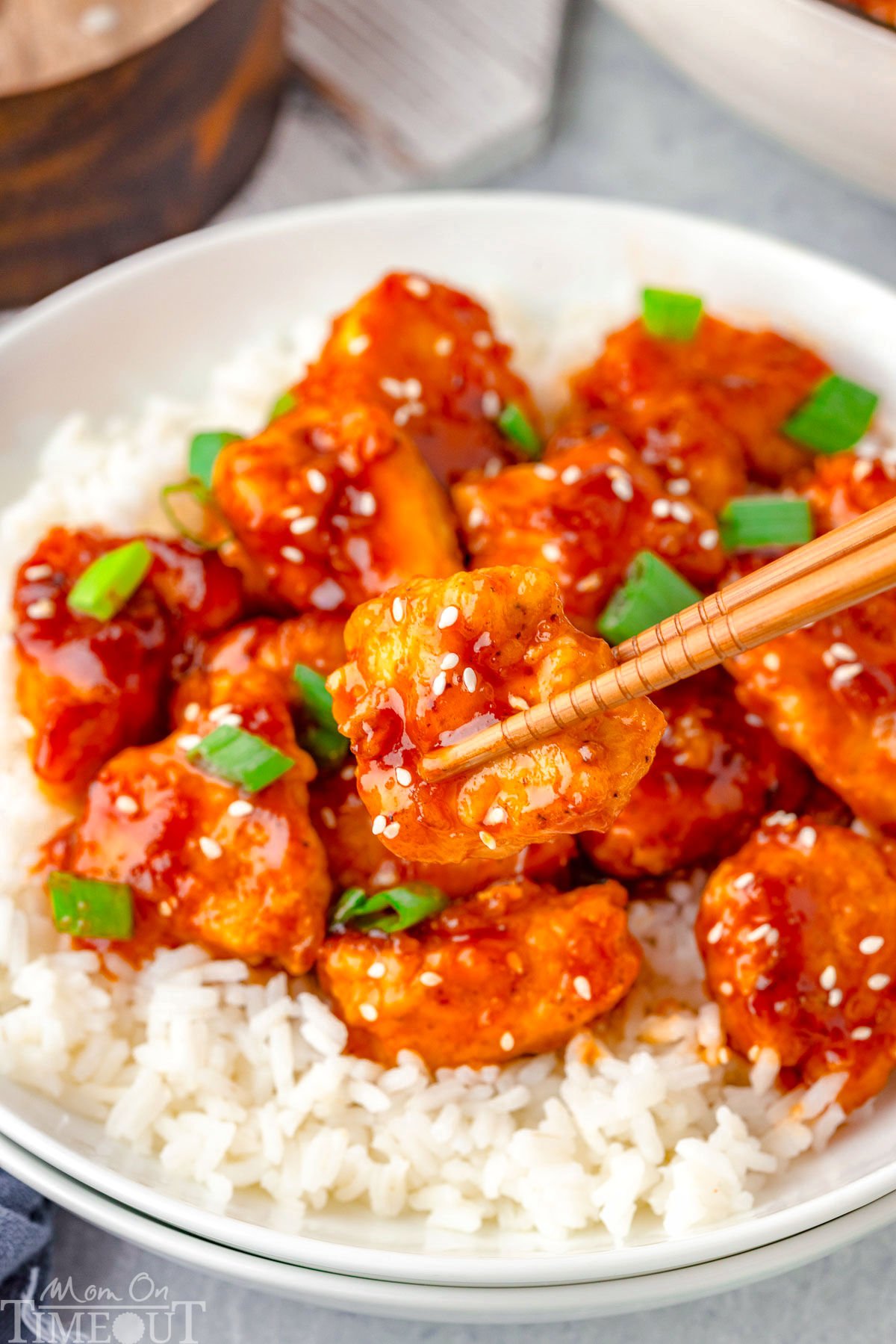 Chopsticks picking up a piece of sesame chicken of a plate with rice and more sesame chicken.