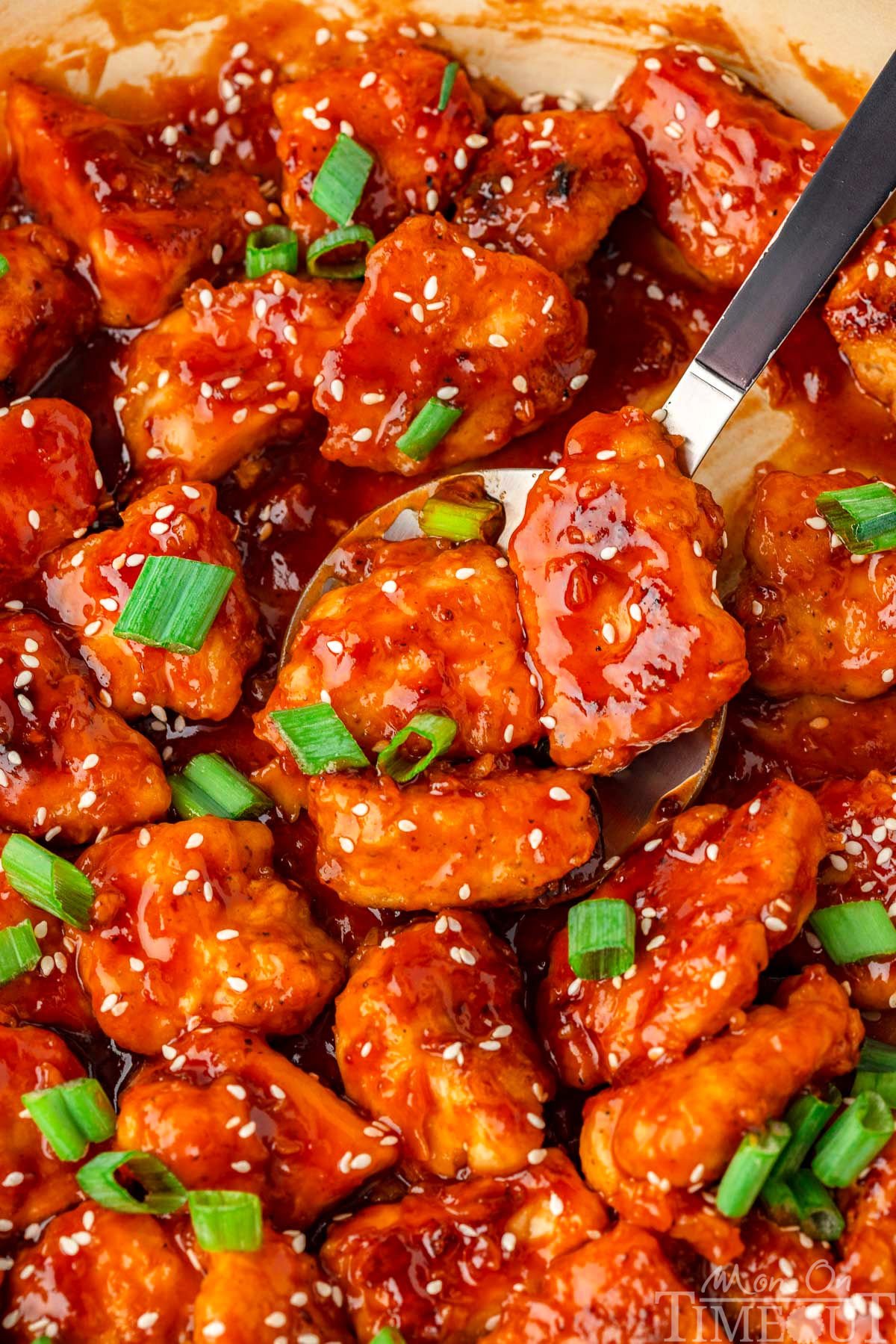 Sesame chicken in sesame chicken sauce in skillet. Spoon is scooping up a few pieces of the chicken.