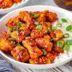 Sesame chicken in a shallow bowl with white rice topped with sesame seeds and green onions. Another bowl can be seen in the background.