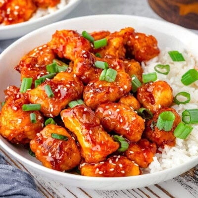 Sesame chicken in a shallow bowl with white rice topped with sesame seeds and green onions. Another bowl can be seen in the background.