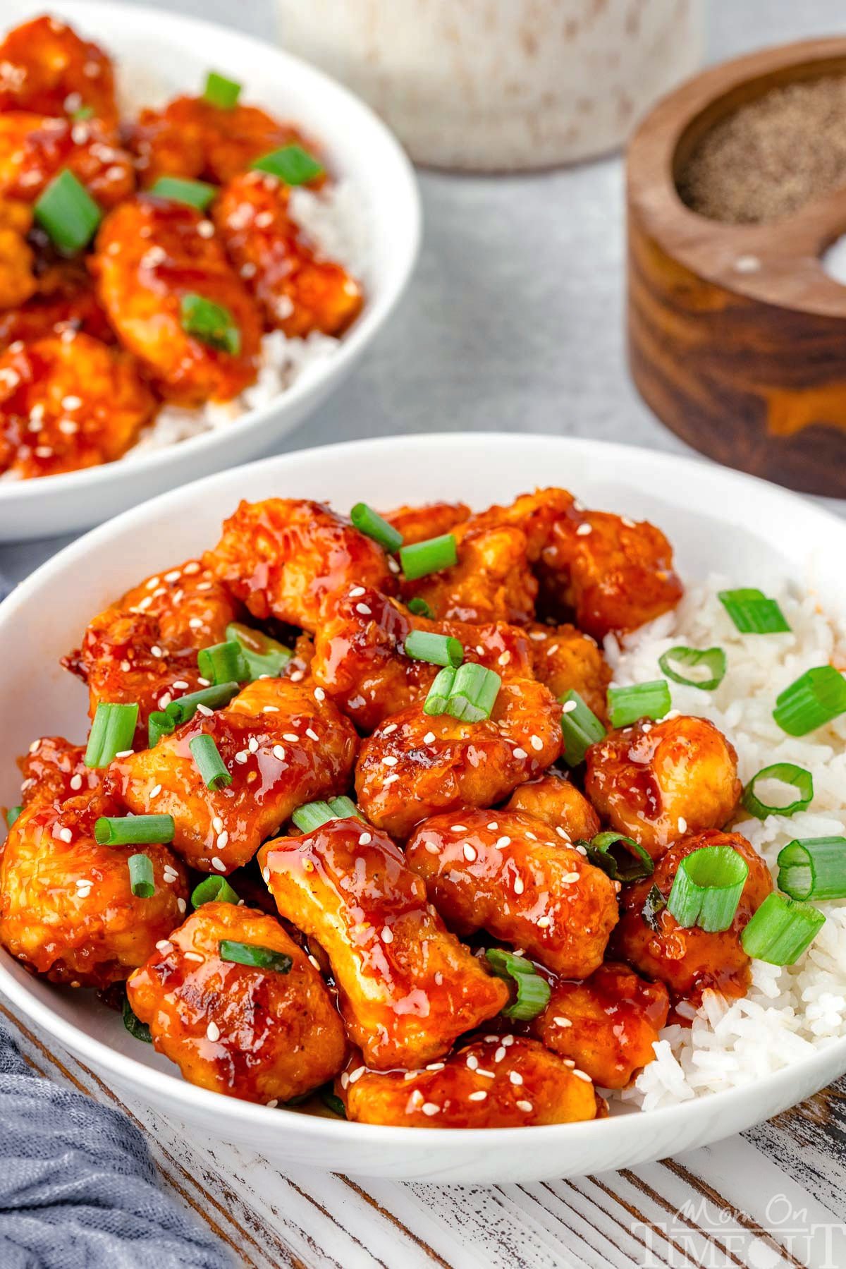 Sesame chicken in a shallow bowl with white rice topped with sesame seeds and green onions. Another bowl can be seen in the background.